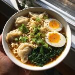 A close-up of a hearty dumpling ramen bowl recipe with noodles, dumplings, soft-boiled eggs, and spinach.