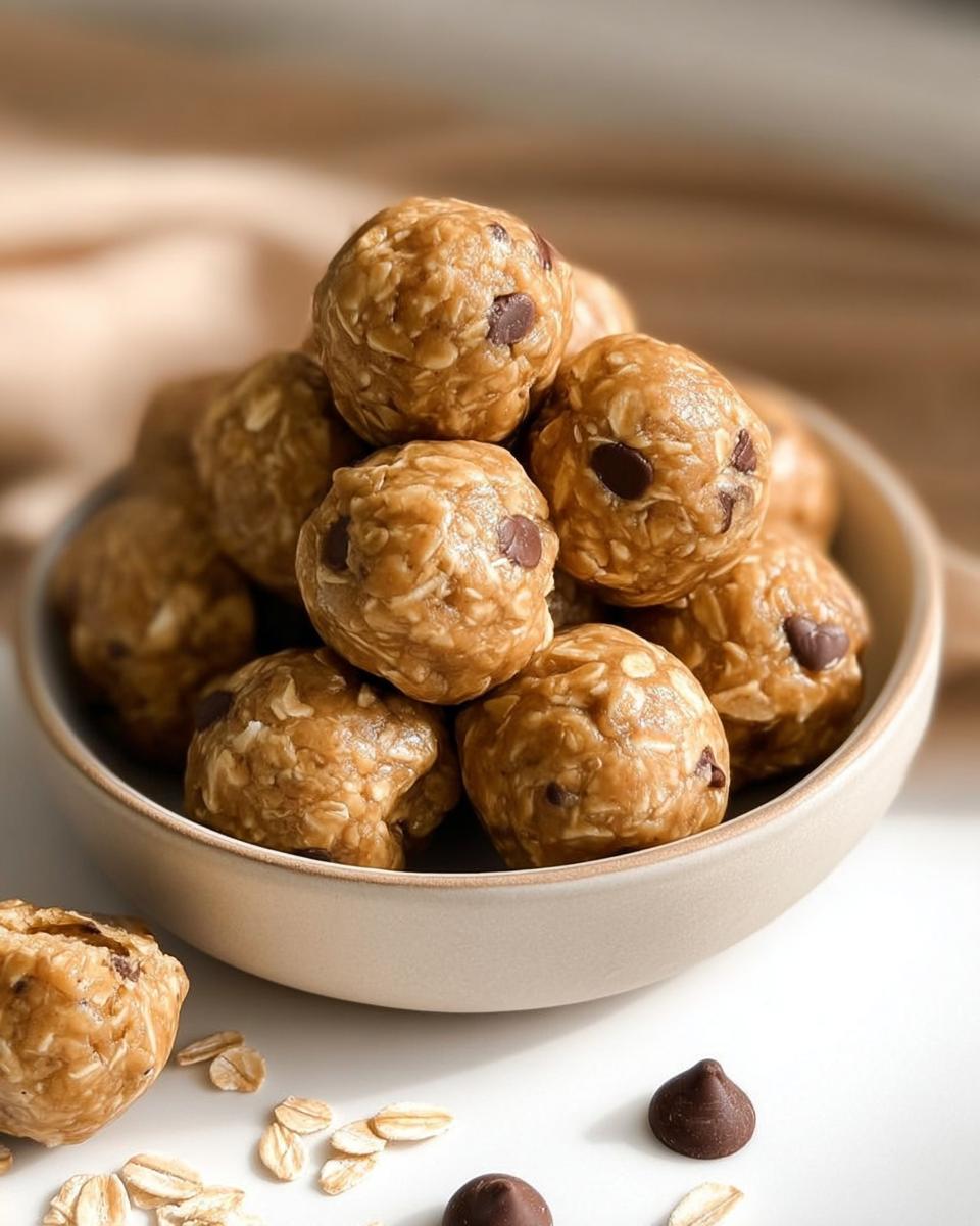 A bowl filled with Easy No Bake Lactation Energy Balls, featuring oats and chocolate chips. Some balls and ingredients are scattered around the bowl.