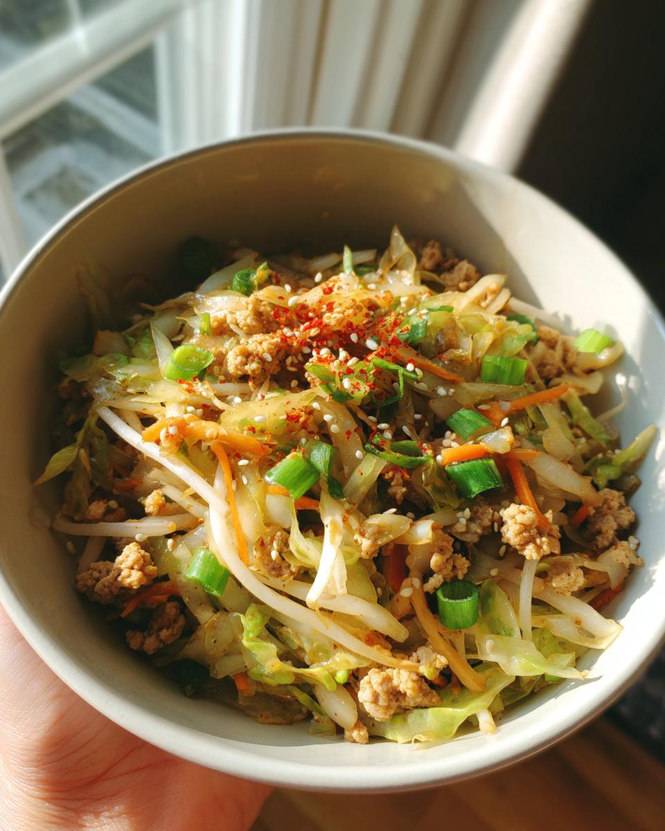 Close-up of a bowl filled with a colorful Egg Roll in a Bowl recipe, featuring ground meat, cabbage, carrots, and bean sprouts, topped with green onions and sesame seeds.