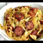 Close-up of a white bowl filled with savory fried cabbage and sausage, garnished with fresh herbs.