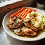 A plate of Garlic Herb Chicken with Mashed Potatoes and steamed carrots, drizzled with sauce and sprinkled with herbs.