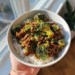 A close-up of a bowl filled with white rice, topped with ground beef and broccoli dinner, garnished with sesame seeds and green onions.