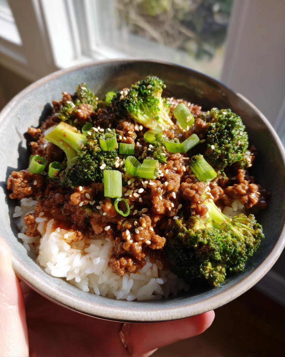 A close-up of a bowl filled with white rice, topped with a savory ground beef and broccoli mixture, garnished with sesame seeds and green onions.