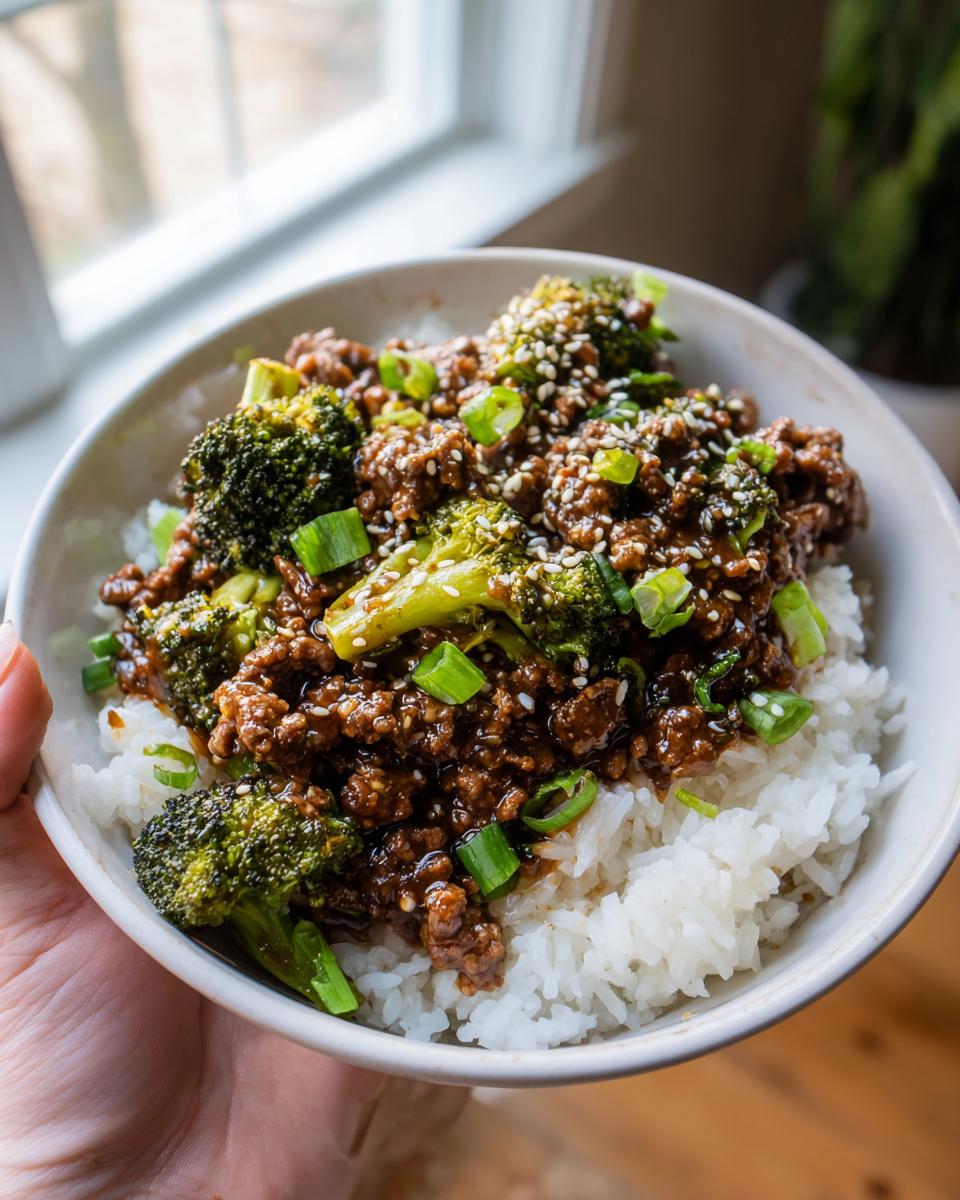 A close-up of a bowl filled with fluffy white rice topped with a savory ground beef and broccoli mixture, garnished with sesame seeds and green onions.