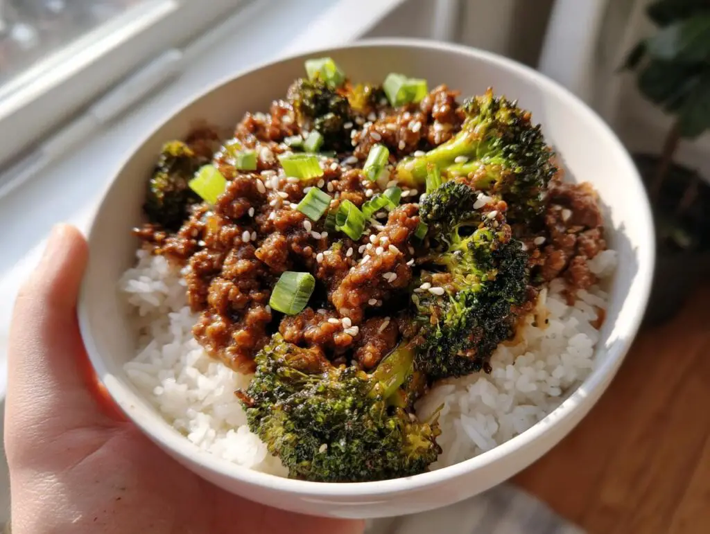 A white bowl filled with white rice, topped with savory ground beef and broccoli, garnished with sesame seeds and green onions.