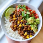 A delicious Ground Beef Taco Bowl filled with rice, seasoned ground beef, black beans, corn, avocado, pico de gallo, and a lime wedge.