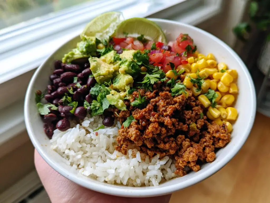 A vibrant Ground Beef Taco Bowl filled with rice, seasoned ground beef, black beans, corn, avocado, salsa, and lime wedges.
