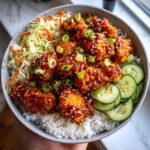 A close-up of a bowl filled with white rice, topped with crispy, glazed Hot Honey Chicken, shredded cabbage, carrots, and sliced cucumbers.
