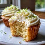 A close-up of a keto coconut lime cupcake with a bite taken out, showing the moist crumb and creamy frosting topped with lime zest.
