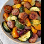 A close-up of a bowl filled with colorful sheet pan sausage and veggies, including zucchini, squash, and red onion.