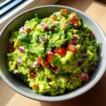 A bowl of chunky simple guacamole recipe, topped with chopped red onion, tomatoes, and cilantro.