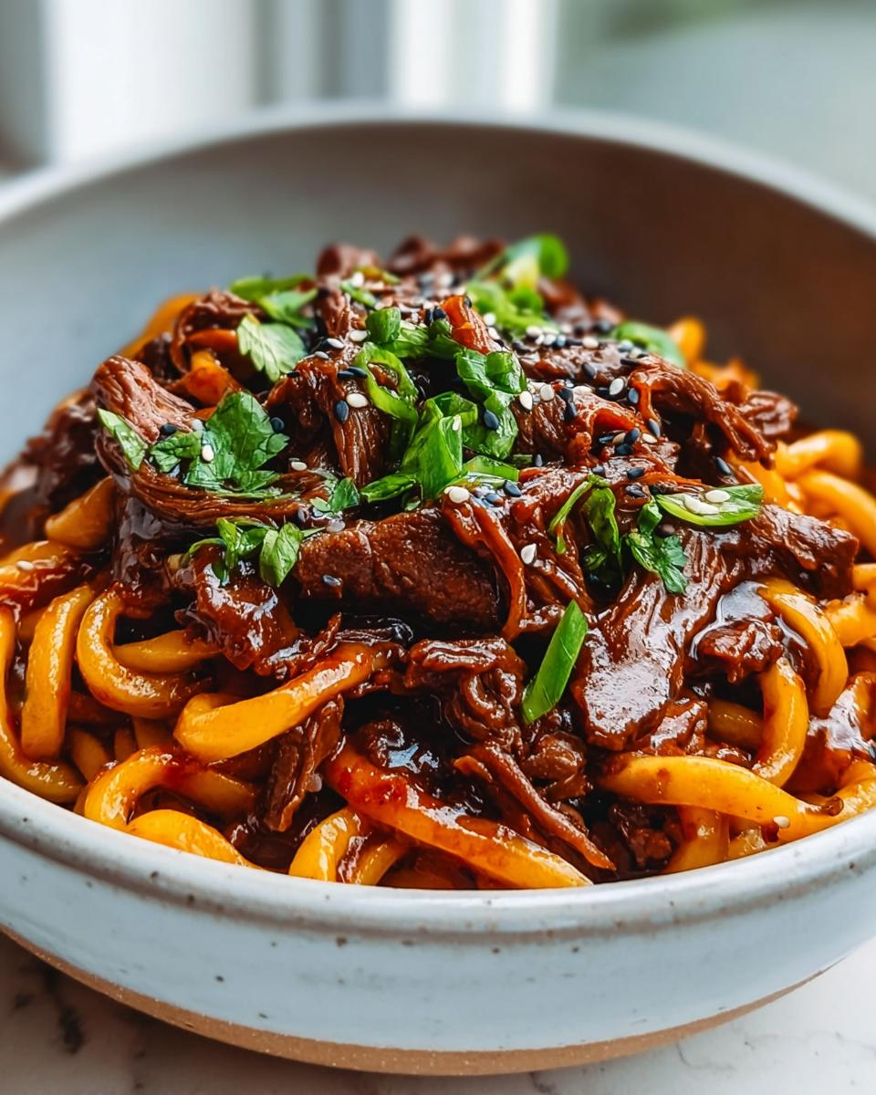 A close-up of Slow Cooker Korean Beef Noodles, featuring tender beef and thick noodles coated in a rich sauce, garnished with sesame seeds and green onions.