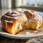 Close-up of two fluffy sourdough cinnamon rolls on a white plate, generously topped with white glaze.