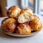 A close-up of a pile of golden-brown sourdough discard pretzel bites, sprinkled with coarse salt. One bite is cut in half, revealing a fluffy interior.