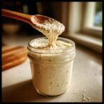 A wooden spoon lifting active sourdough starter from a glass jar, showing its bubbly texture.