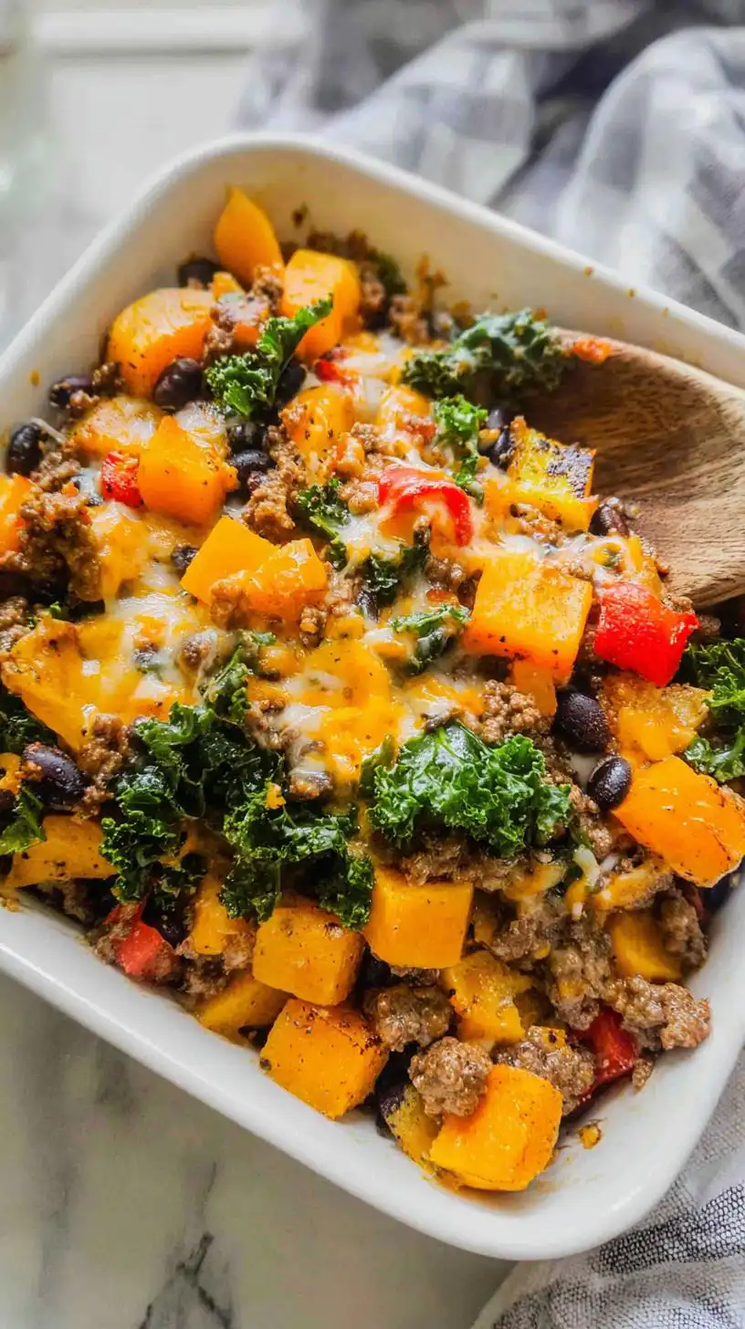 A close-up of a baked Ground Beef Butternut Squash Casserole in a white dish, topped with melted cheese and kale.