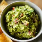 A close-up of fresh Mary Berry Guacamole in a bowl, with chunks of tomato and coriander, served with tortilla chips.