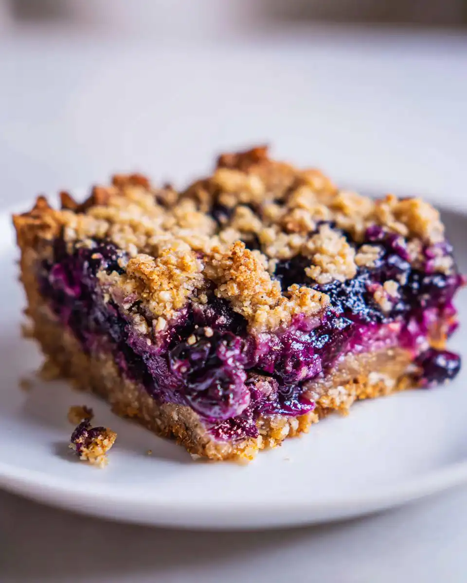 Close-up of a wholesome blueberry oatmeal bar slice on a white plate, showing a crumb topping and juicy blueberries.