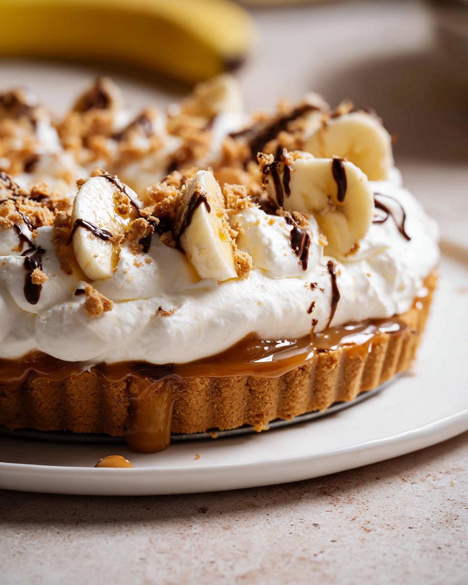 Close-up of a delicious Banoffee Tart Recipe, Mary Berry inspired, topped with whipped cream, sliced bananas, chocolate drizzle, and biscuit crumbs.
