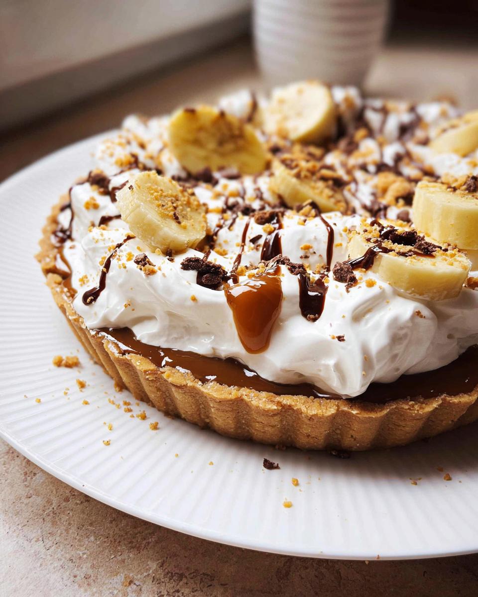 A close-up of a delicious Banoffee Tart Recipe, inspired by Mary Berry, topped with whipped cream, banana slices, chocolate drizzle, and biscuit crumbs.