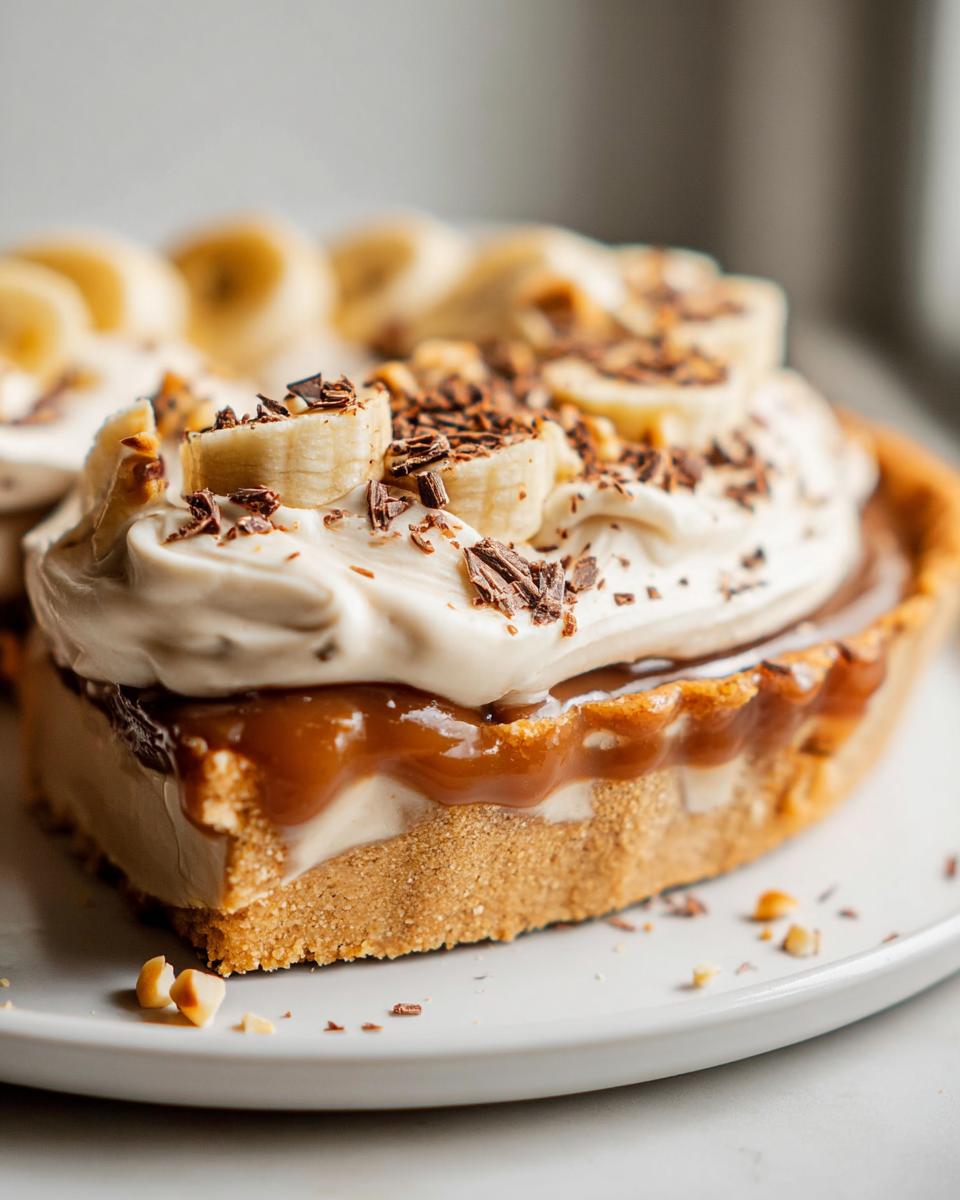 A decadent slice of Mary Berry Banoffee Pie, featuring layers of biscuit base, toffee, banana, and whipped cream, topped with chocolate shavings and nuts.