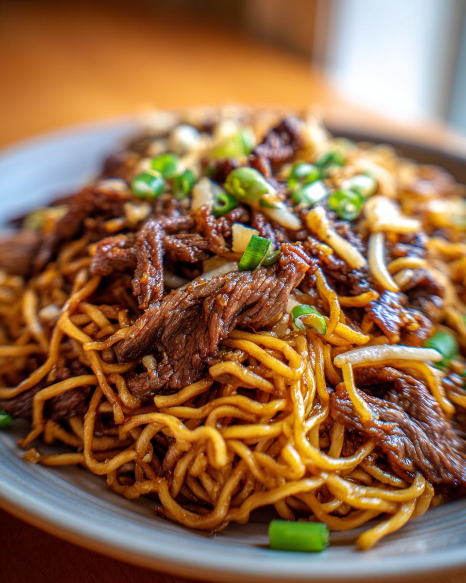 Close-up of a plate of Mongolian Beef Noodles, featuring tender beef strips and golden noodles, garnished with green onions.